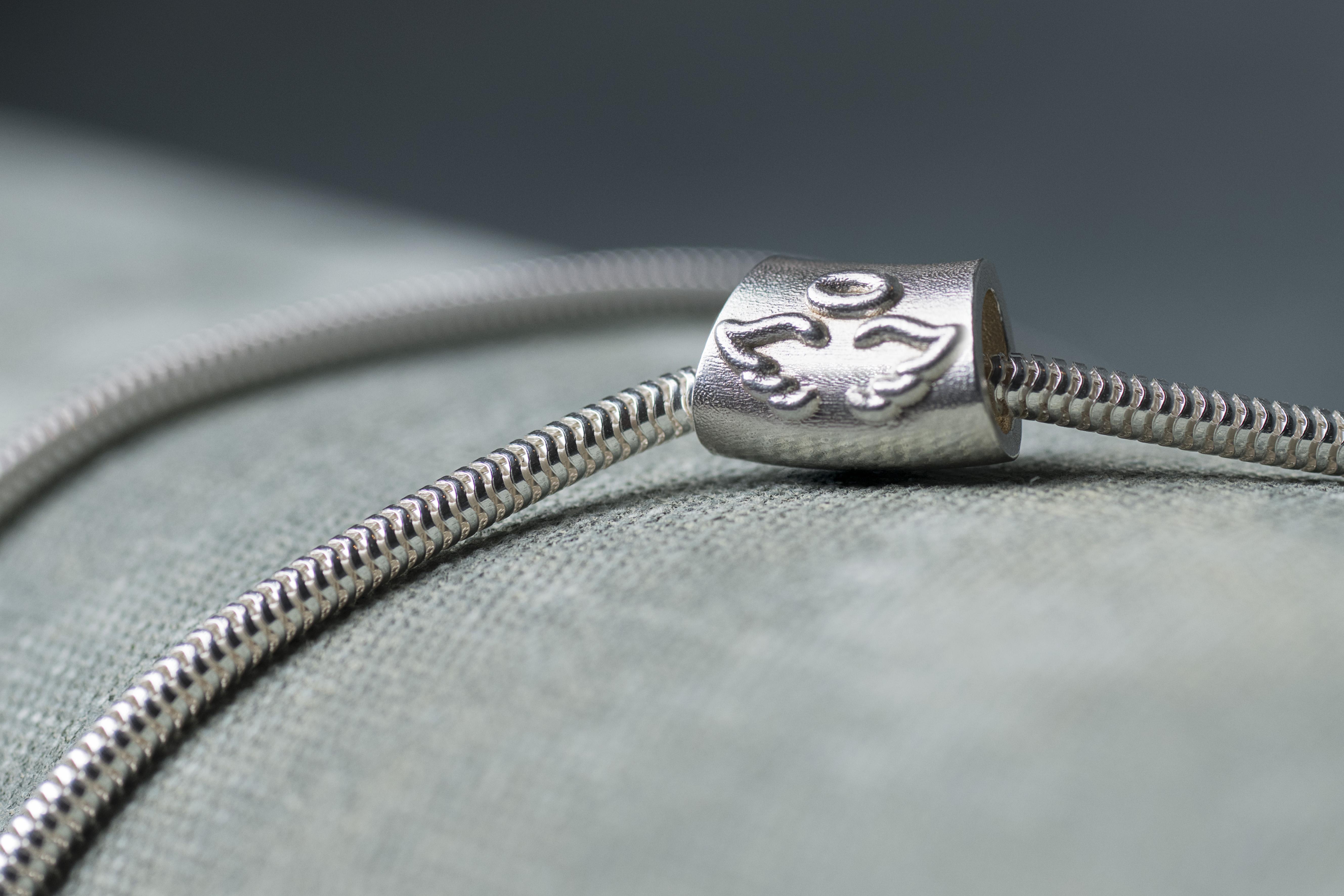 Close-up of the silver Angel Baby Charm with raised angel wings and a halo, threaded onto a silver snake chain on a pale grey surface