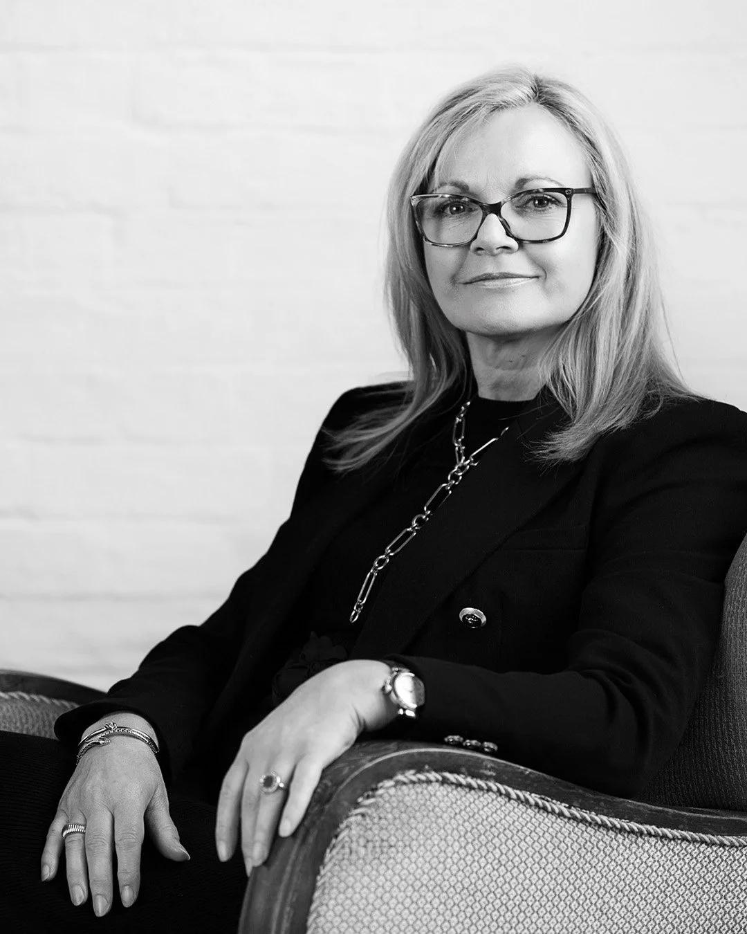 Black-and-white seated portrait of GC&DC Chair Judith Lockwood wearing glasses, a dark tailored jacket and jewellery
