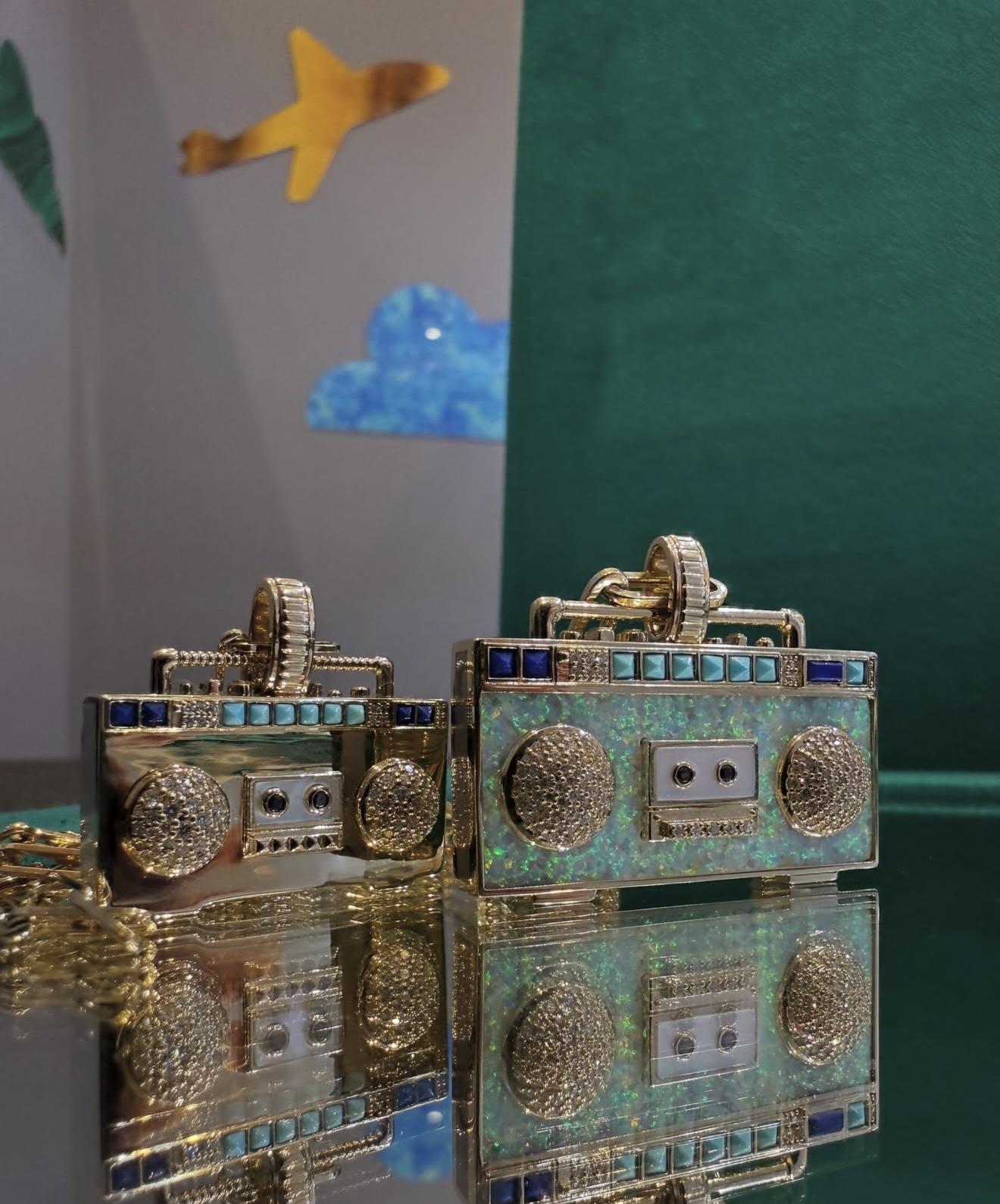 Two NeverNoT Turn It Up boombox pendants in yellow gold featuring diamonds, opal and blue gemstones displayed on a reflective surface