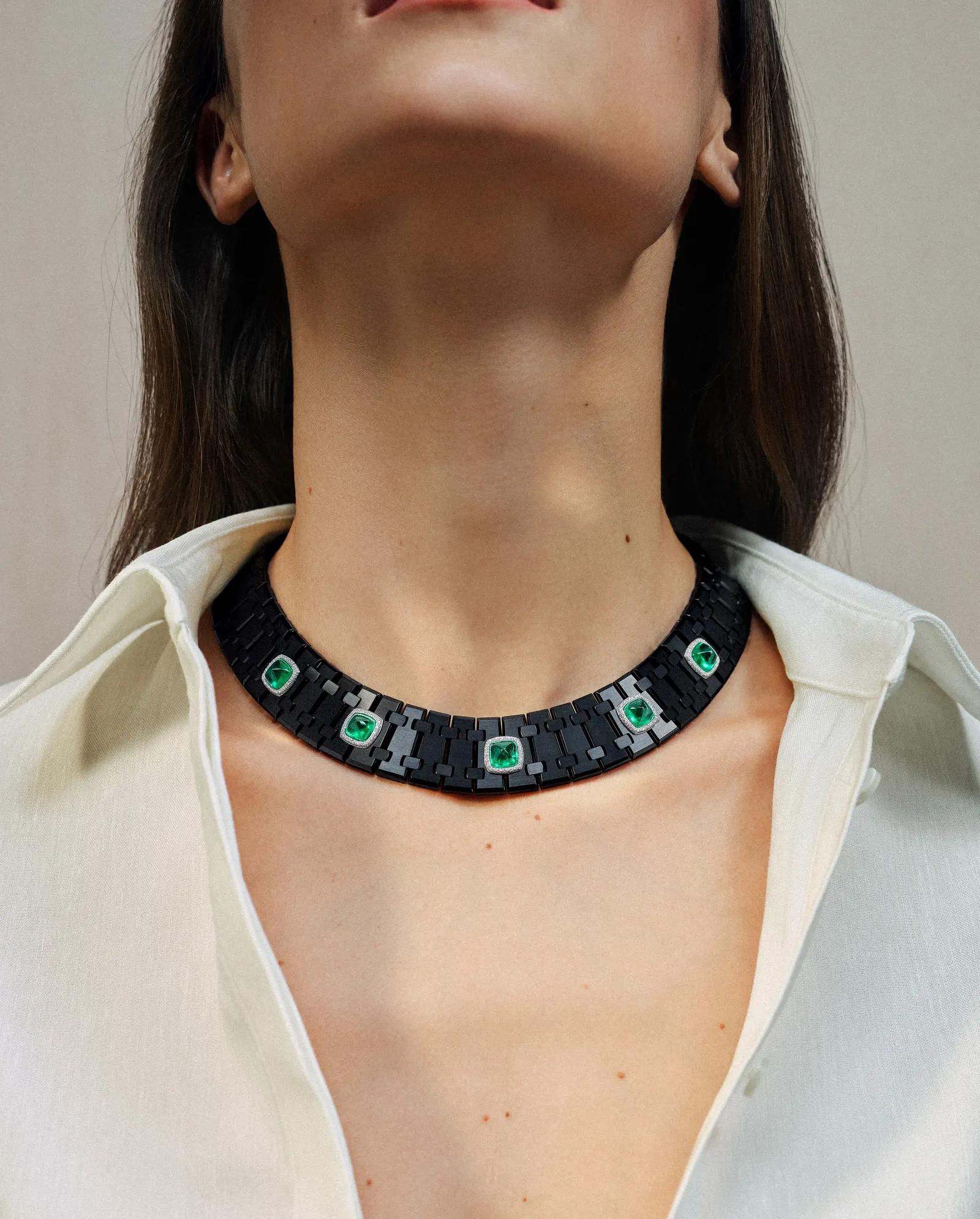 Model wearing a wide black articulated Audemars Piguet necklace set with five vivid green sugarloaf emeralds surrounded by white diamonds