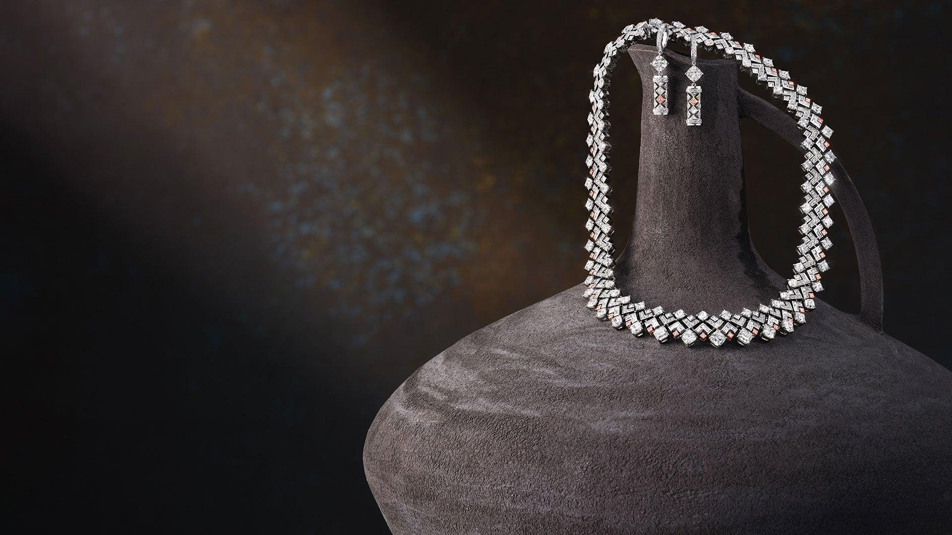 Geometric white-diamond necklace and drop earrings with small pink diamond accents, displayed on a sculptural dark-grey stand.