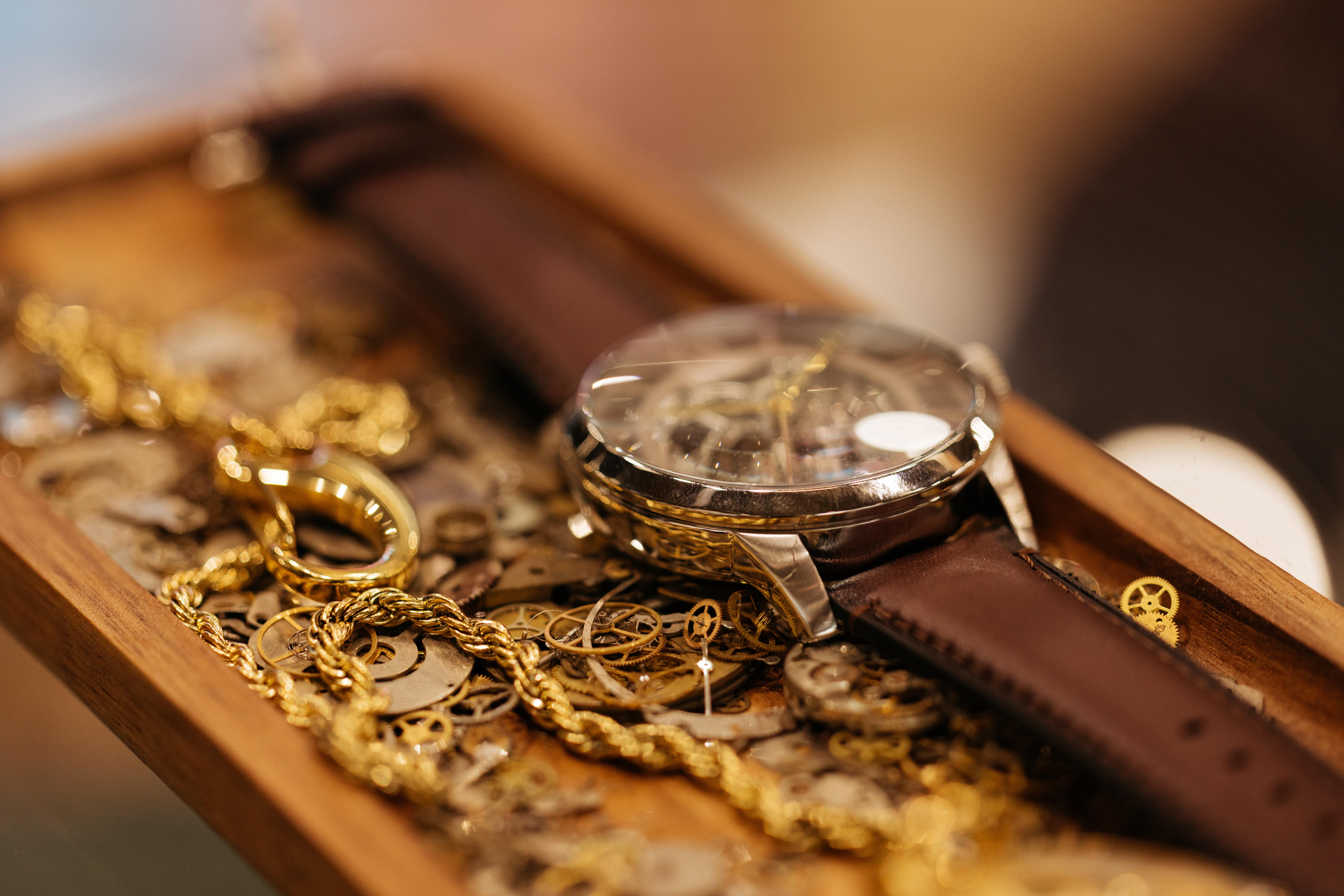 Close-up of a vintage wristwatch with a brown leather strap, a gold chain and small watchmaking gears arranged inside a wooden tray.
